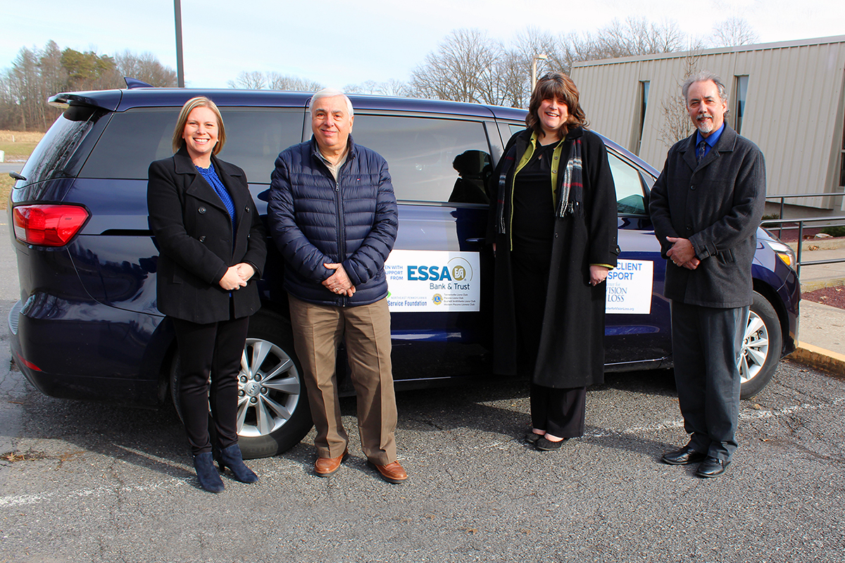 Four people standing next to a new guided transport vehicle, a dark blue mini van