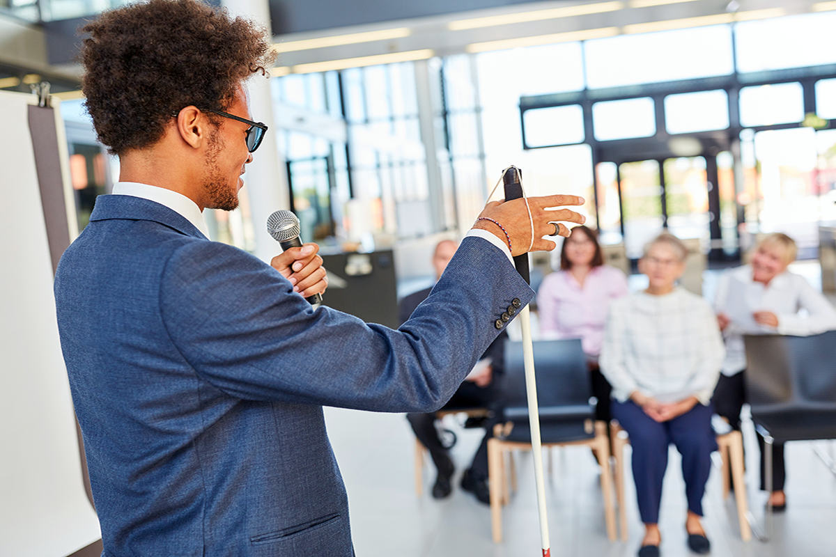 A man holding a white cane and a microphone speaking before an audience