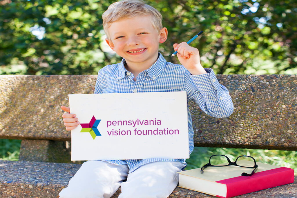 Photo of a young boy sitting on a park bench next to a pair of glasses and a book while holding a sign with the Pennsylvania Vision Foundation logo