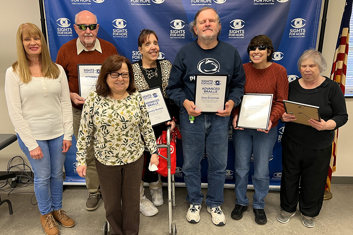 Advanced Braille class graduates, their instructor, and their instructors in front of a Sights for Hope step-and-repeat banner