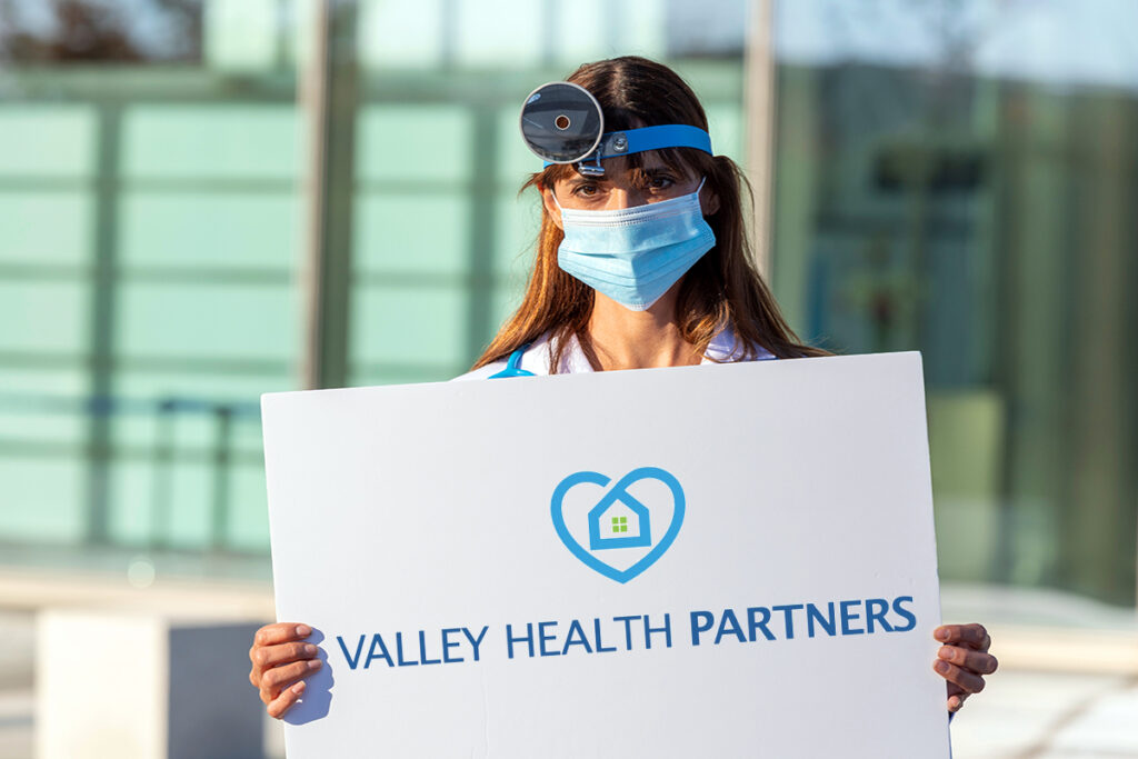 Phot of a female doctor wearing a white coat and mask holding a white sign with the Valley Health Partners logo