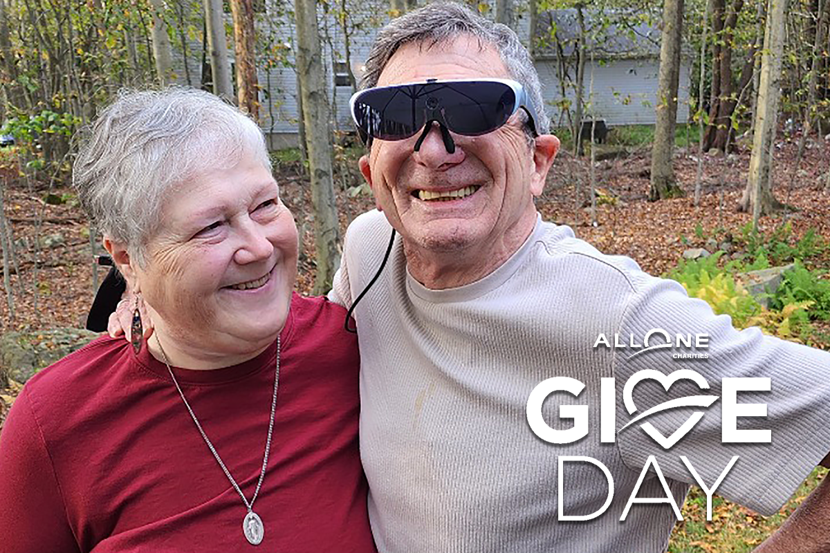 The AllOne Charities Give Day logo imposed upon a photo of Sights for Hope client Ray, who is wearing a special piece of assistive technology to help him see more of the world, outdoors with his wife, Joan.