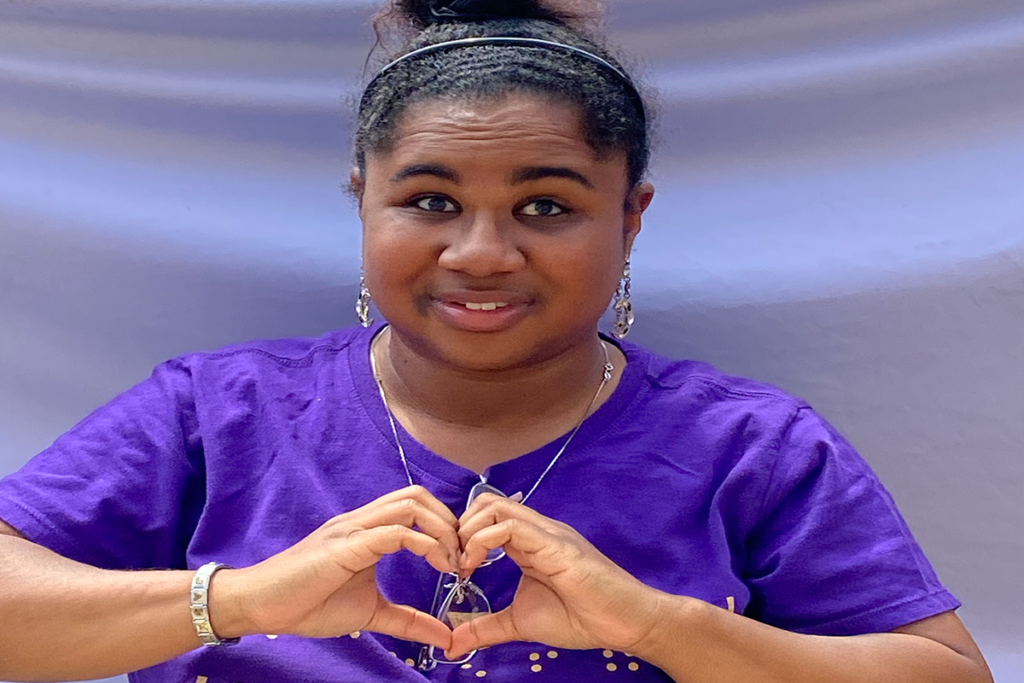 Sights for Hope client Jasmyn makes a heart symbol with her hands; she is wearing and dark purple shirt and is in front of a light purple backdrop