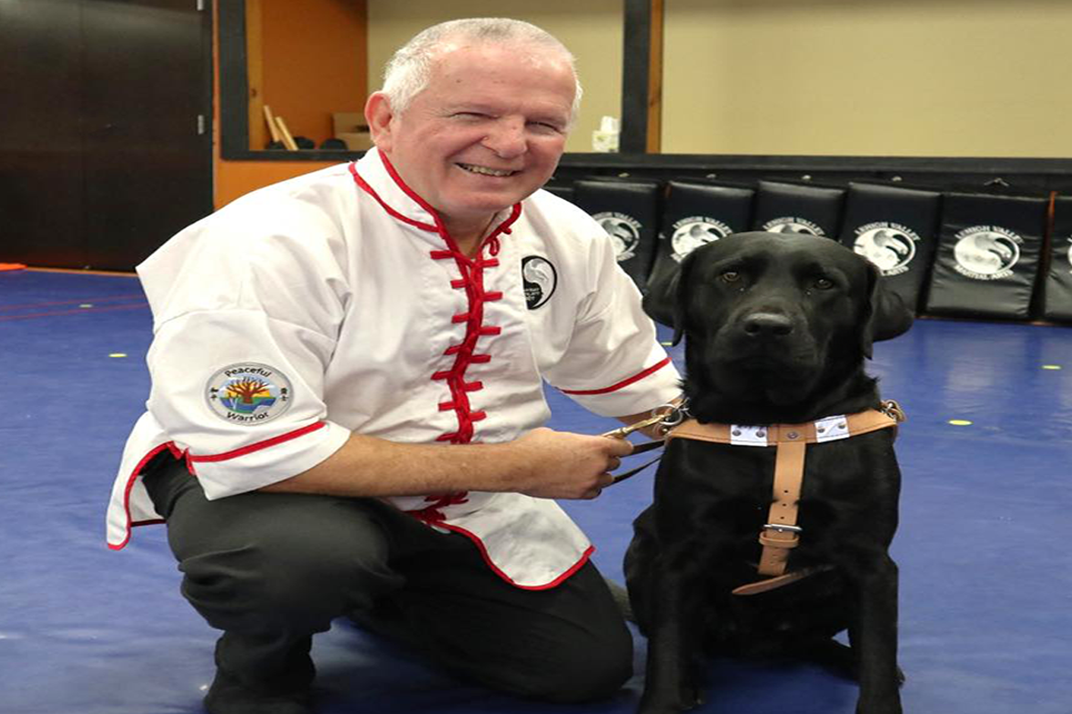 Paul Miller in a white martial arts uniform and black pants kneeling next to his guide dog, Felton, a black Labrador