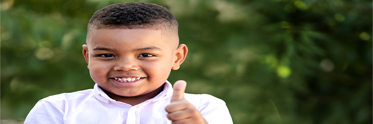 Smiling child wearing a white shirt gives a thumbs-up against a blurred green background, conveying positivity and confidence.