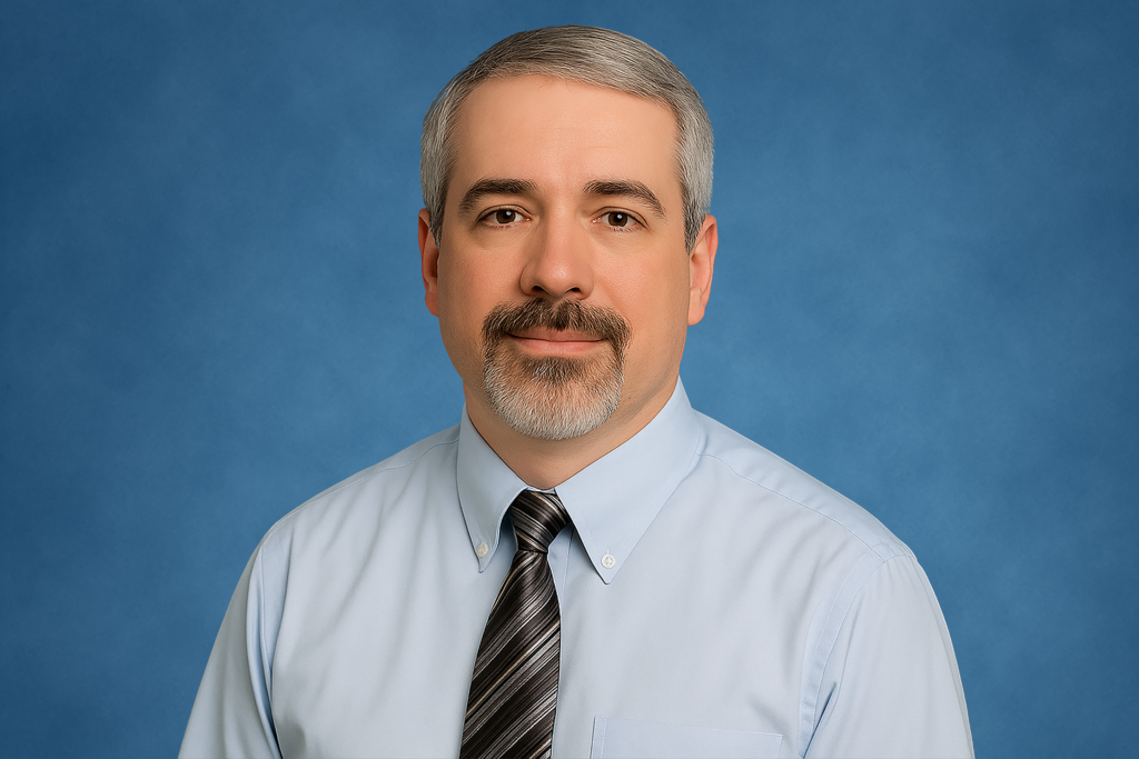 A man with short gray hair and a beard wearing a light blue shirt and striped tie. He is smiling slightly against a solid blue background. Professional tone.