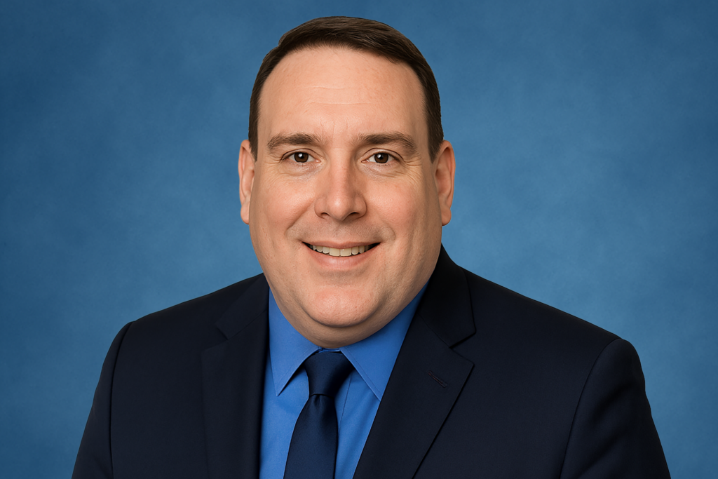 A man in a black suit with a blue shirt and tie smiles against a solid blue background. The image conveys a professional and approachable tone.