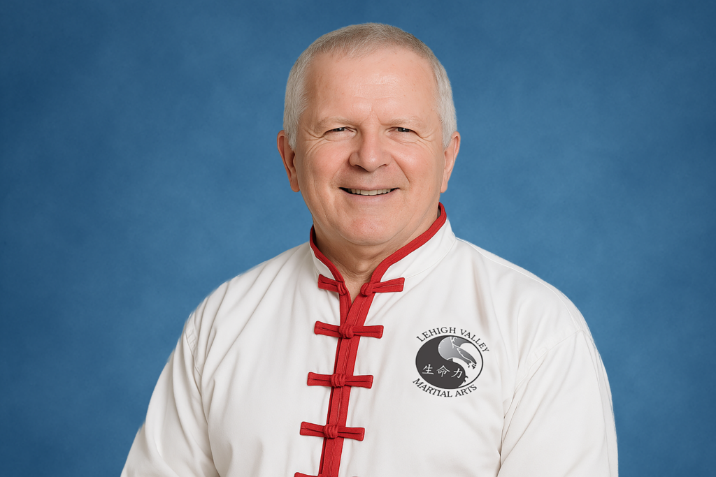 Elderly man smiling, wearing a white martial arts uniform with red trim and a "Lehigh Valley Martial Arts" logo, against a blue background.