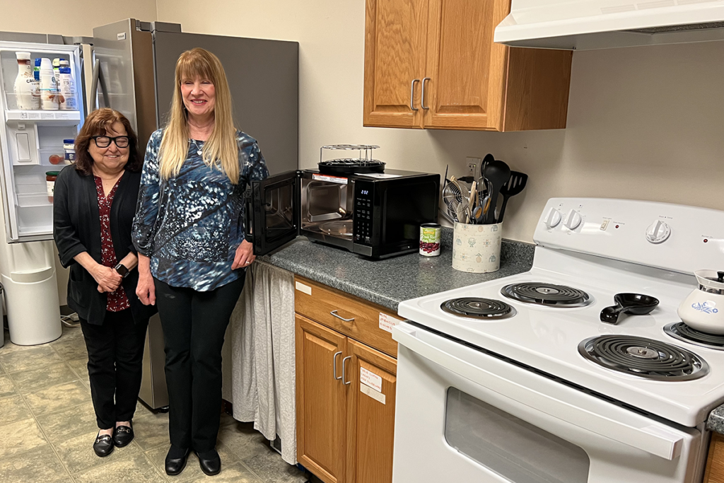 Dianne Michels and Rita Lang of Sights for Hope and the Lehigh Valley Council of the Blind smile inside Sights for Hope's Lehigh Valley Services Center kitchen. The scene features an open fridge, a microwave with its door open, wooden cabinets, and an electric stovetop.