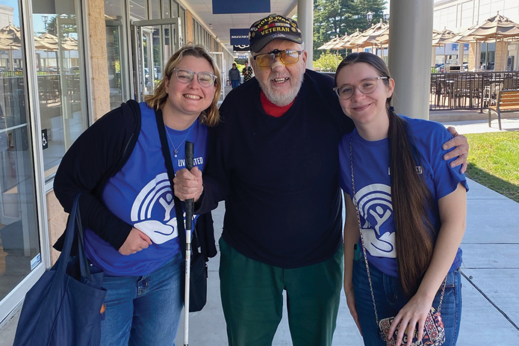 A beaming man wearing a "Veteran" cap is joyfully embraced by two women in blue "Live United" shirts. They stand outside, exuding warmth and camaraderie.