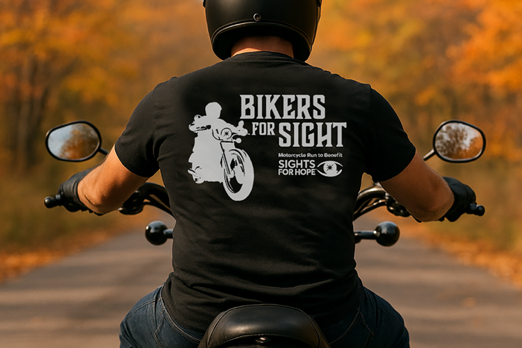 A biker with a bald head, black helmet, and black T-shirt that reads "Bikers for Sight" and "Motorcycle Run to Benefit Sights for Hope," rides through a tree-lined road in autumn, conveying a sense of freedom and purpose. Generated with artificial intelligence.