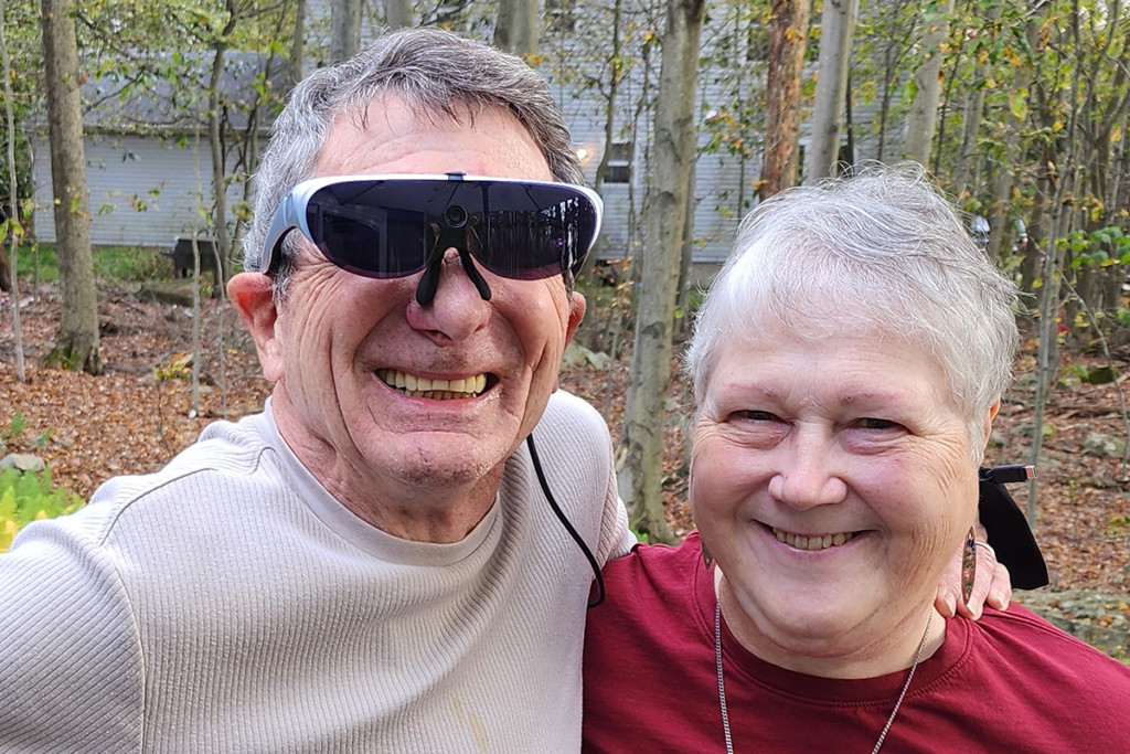 An older couple smiles warmly in a wooded area. The man is wearing an assistive device for his vision. Autumn leaves set a cozy tone.