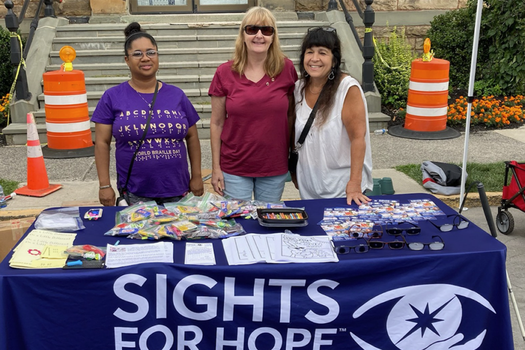 Three women – who are Sights for Hope clients and members of the Monroe Sights for Hope Lions Club – stand behind a table covered by a purple tablecloth and various items. They are smiling and appear welcoming. The background features steps and orange cones.