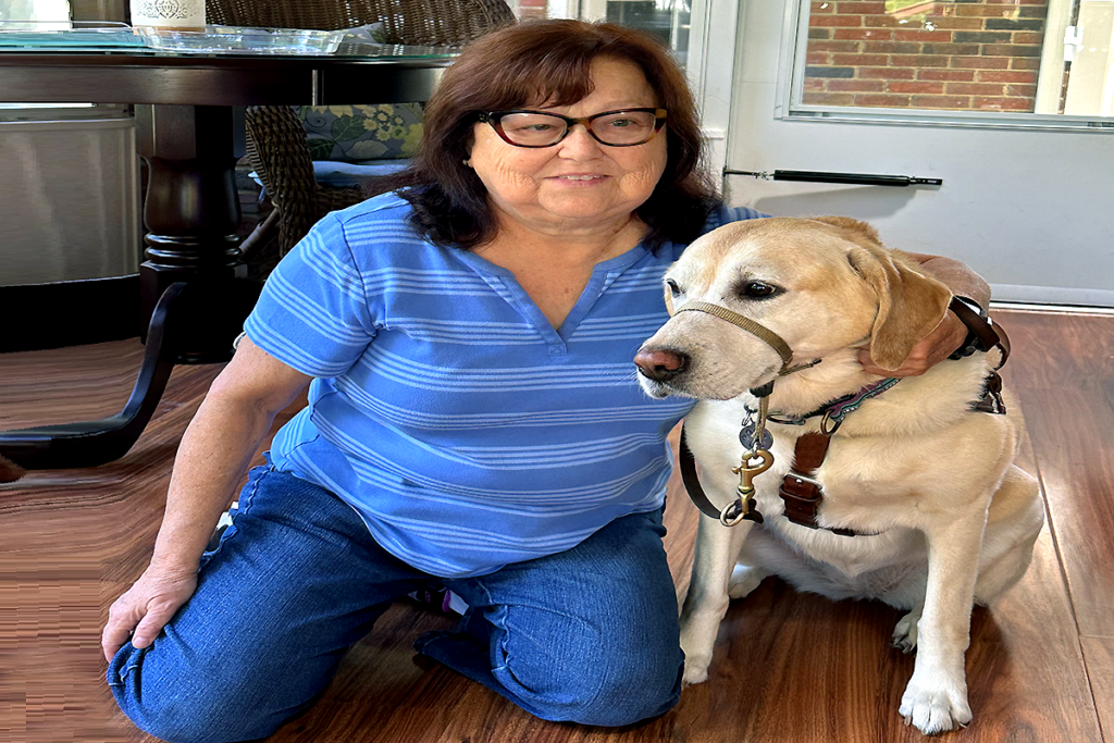 Dianne Michels, in a blue striped shirt, kneeling on a wooden floor next to her service dog, Sammie, a yellow Labrador-Golden Retriever cross who is wearing a harness.