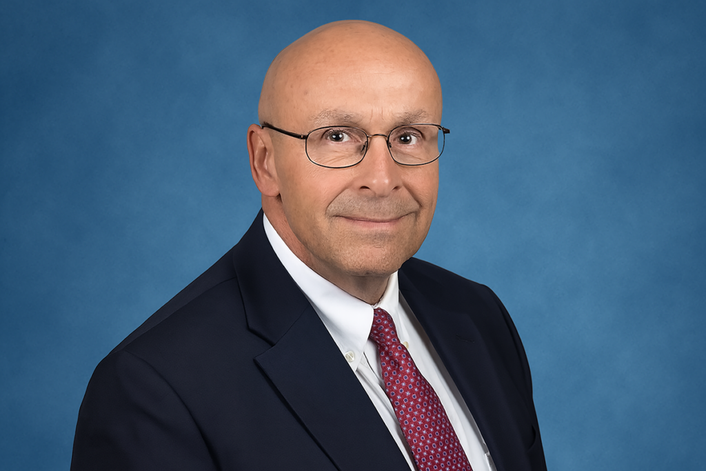 A man with a bald head and glasses smiling warmly, wearing a dark suit, white shirt, and red patterned tie against a plain blue background, conveys professionalism. Generated with artificial intelligence.