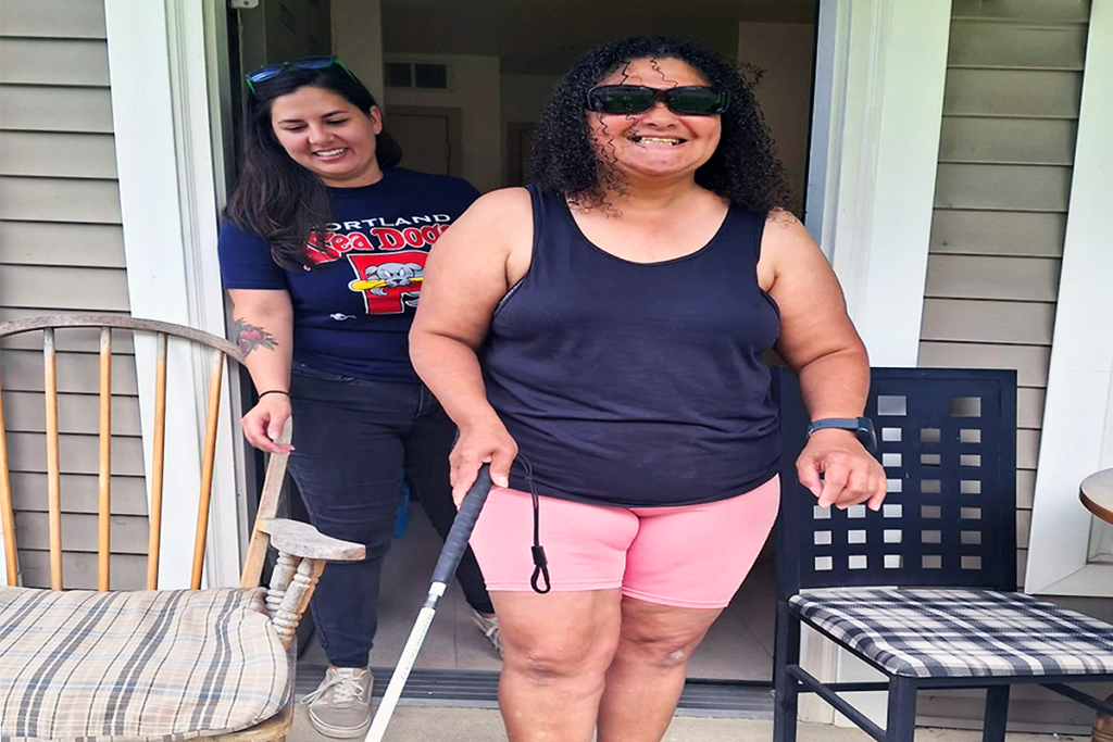 Two women stand in a doorway, smiling. The woman in front uses a white cane and wears sunglasses. The scene conveys a joyful, supportive atmosphere. The woman in the foreground is a Sights for Hope client. The woman behind her is Sights for Hope O&M instructor Tesia Nasehi.