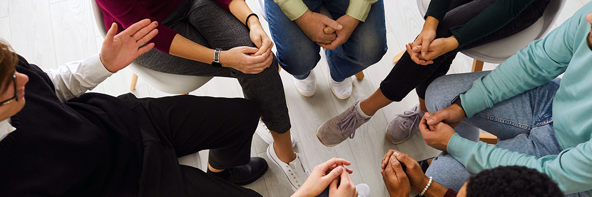 A diverse group sits in a circle, focusing intently, suggesting discussion or support. Hands show engagement, conveying a sense of connection and community.