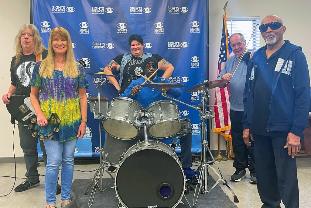A diverse group of five people poses with musical instruments, a drum set in the center. They stand smiling in front of an event banner and a U.S. flag.