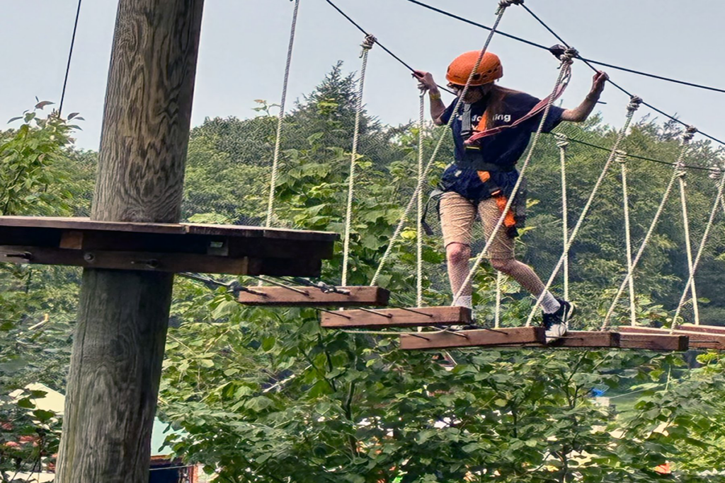 A Transitions program client wearing a helmet and harness navigates a rope bridge in an adventure park. Background features lush green trees, conveying excitement and focus.