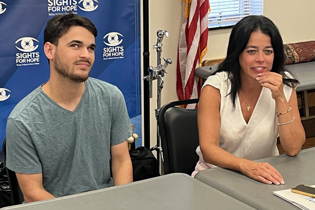 Two people sit at a table in a room, one in a gray shirt and one in a white blouse. A Sights for Hope banner and a U.S. flag are in the background.