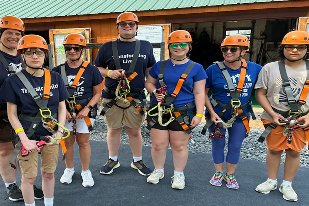 A group of teens and pre-teens wearing orange helmets and harnesses stand side by side, smiling. They are on a zip-lining adventure, exuding excitement and teamwork.