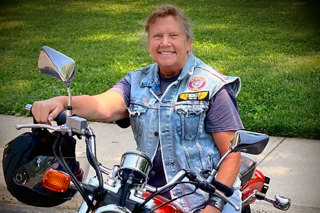 A woman smiling while sitting on a motorcycle in a park. The woman is Sights for Hope client Wendy Smentkowski, who inspired the Bikers for Sight event. She is wearing a denim vest with patches.