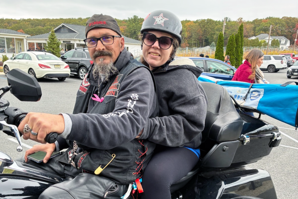 A couple on a motorcycle; the man, wearing a bandana and sunglasses, confidently holds the handlebars. The woman, in a helmet and hoodie, smiles warmly. Background shows parked cars and autumn trees under a cloudy sky.