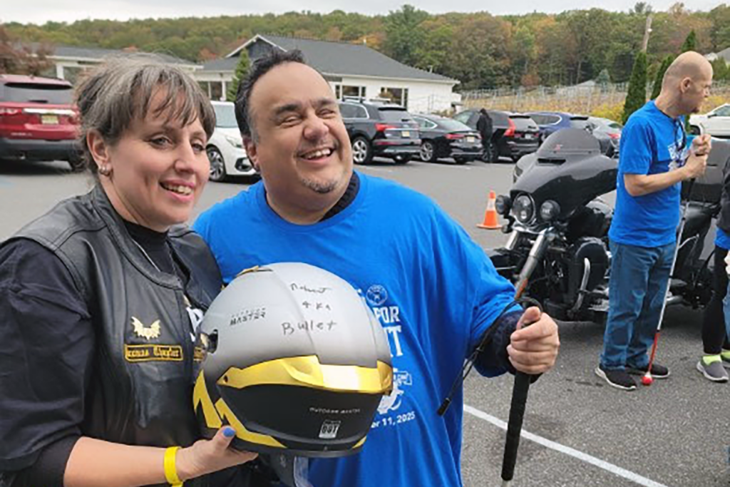 A joyful man and woman stand together, both smiling. The man wears a blue shirt, holding a white cane, and the woman wears a black leather vest, holding a helmet. Two motorcycles and several cars are parked in the background. The scene is outdoors, with overcast skies and trees visible.
