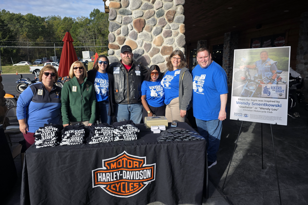 A group of seven people stand smiling behind a table covered with Bikers for Sight shirts, beside a large event poster. The setting is sunny and outdoors, with motorcycles in the background, conveying a friendly and enthusiastic tone.