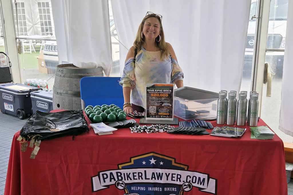 Smiling woman stands behind a table covered in promotional items, including bottles and keychains. A banner reads "PABikerLawyer.com" promoting legal services for bikers.