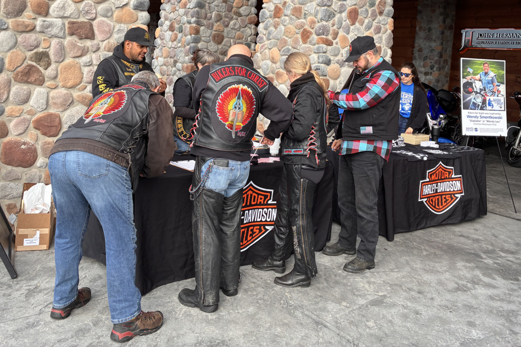 A group of bikers wearing leather vests, including "Bikers for Christ" logos, gather around the Bikers for Sight registration table. The mood is engaged and communal.