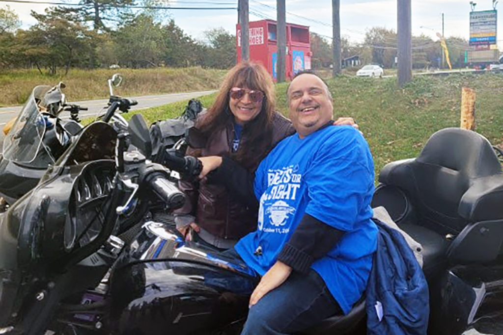 A woman in sunglasses stands beside and a man in a bright blue Sights for Hope shirt, who is sitting on a black motorcycle.
