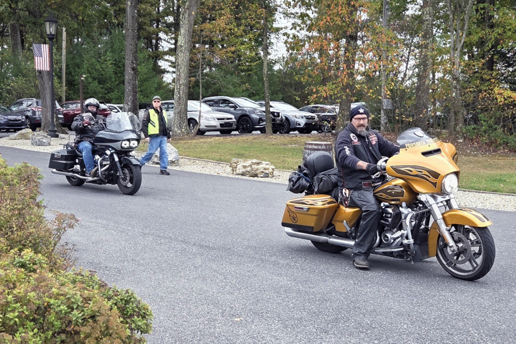 Two motorcyclists ride on a tree-lined road, with a golden and a black motorcycle. Parked cars and a person in a safety vest are in the background.