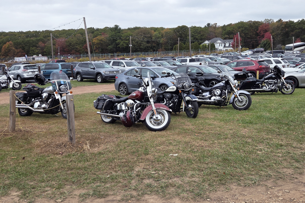 Several motorcycles are parked on grass next to a crowded parking lot filled with cars. A scenic background of trees with autumn foliage is visible.