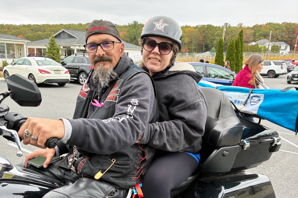 A man and woman on a motorcycle in a parking lot. Both wear helmets and sunglasses, smiling. Background includes parked cars, trees, and people.