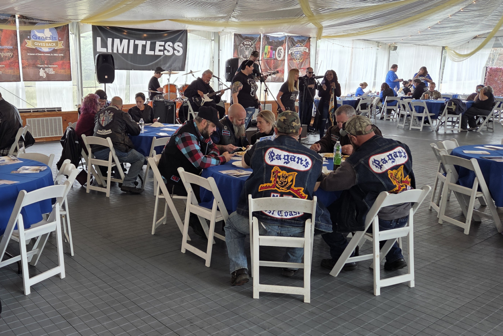 A community event in a hall with people seated at round tables covered in blue cloth. The Sights for Hope client-led band, Limitless, performs on stage, creating a lively atmosphere. Attendees wear jackets with vibrant patches.