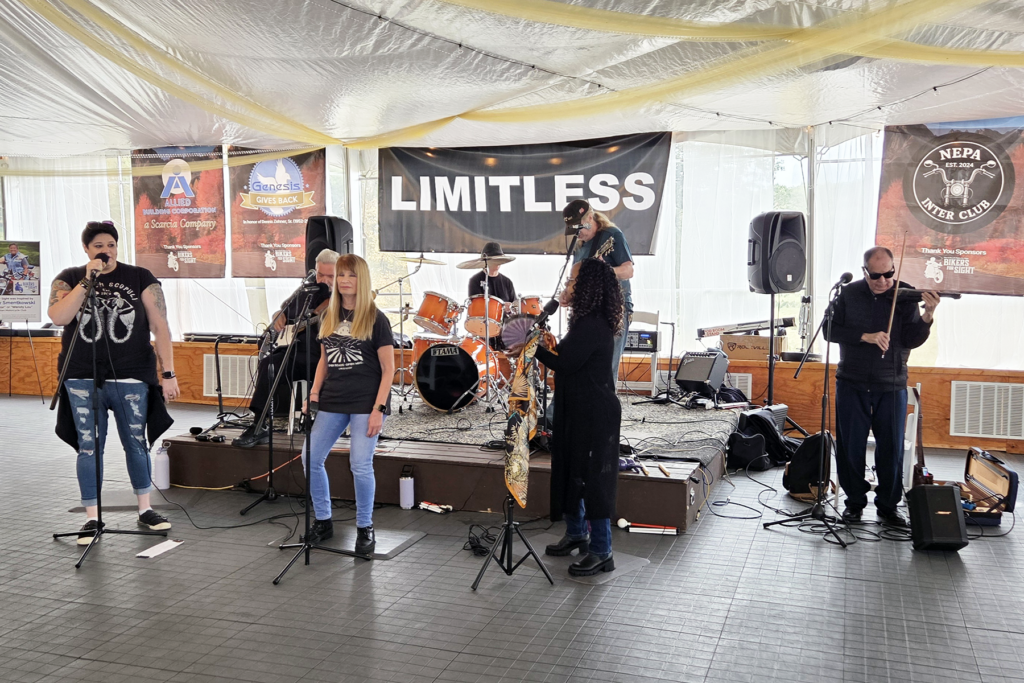 The Sights for Hope client-led band, Limitless, performs on a stage with three vocalists, a guitarist, drummer, saxophonist, and violinist. The setting conveys an energetic and lively atmosphere.
