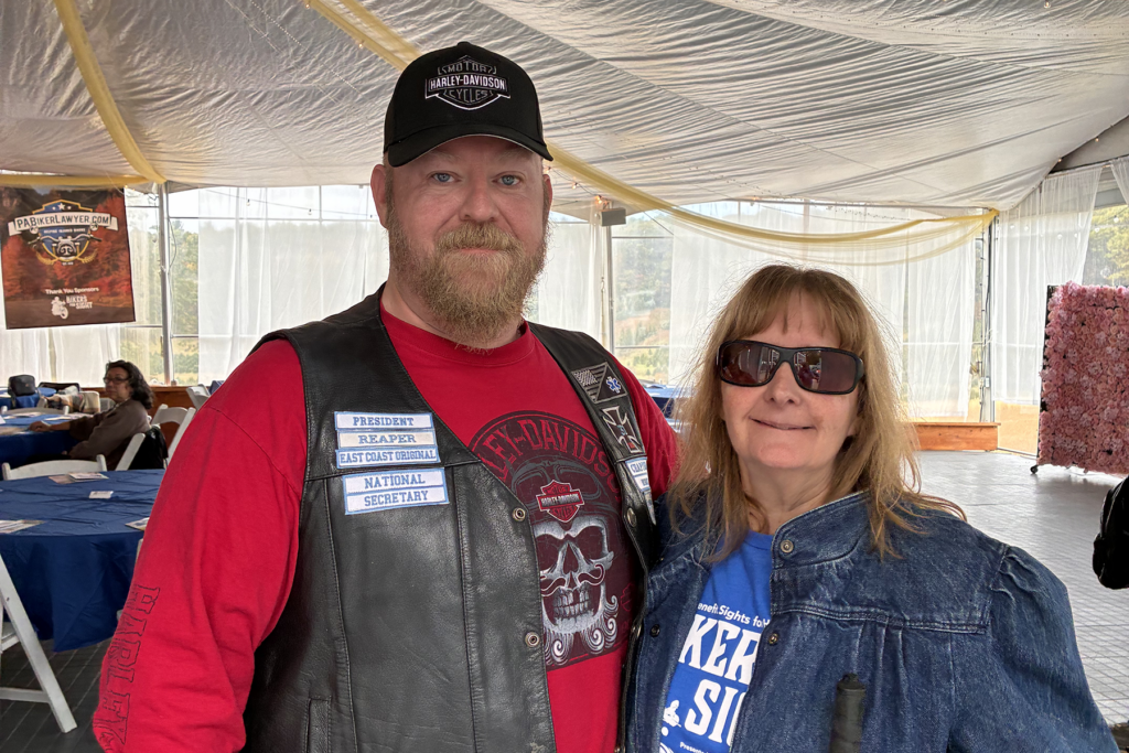 A man in a leather vest and cap stands beside a woman in sunglasses and denim. They smile under a tent with tables behind them.