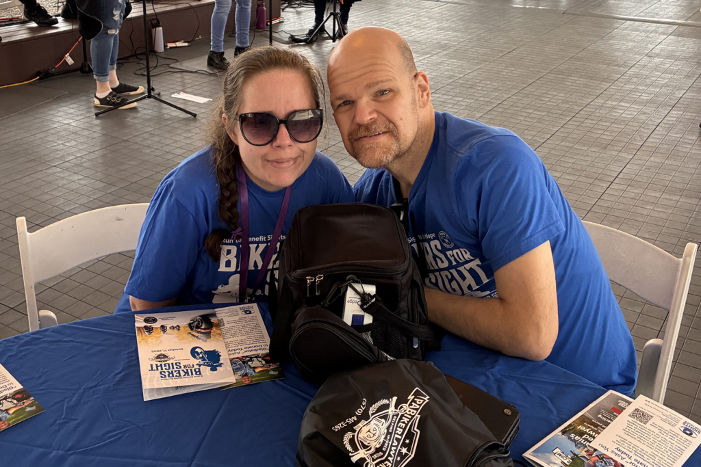 A man and woman wearing matching blue t-shirts sit together at a table. Both are smiling, creating a warm and friendly scene.