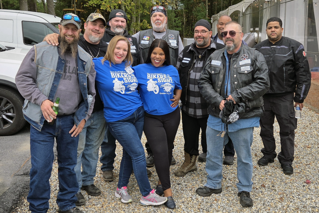 A group of smiling men wearing biker attire pose outdoors with two women in blue Bikers for Sight event shirts. The atmosphere is cheerful and friendly.