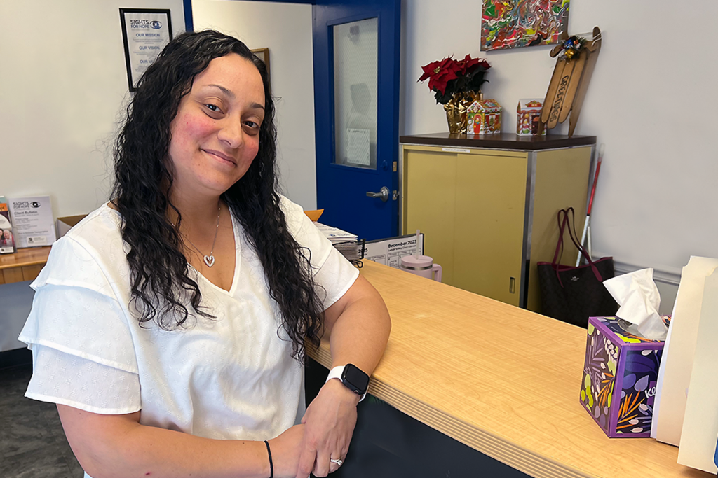 A woman with long dark hair and a heart necklace smiles warmly, leaning on a counter. Behind her are a blue door and a tissue box.