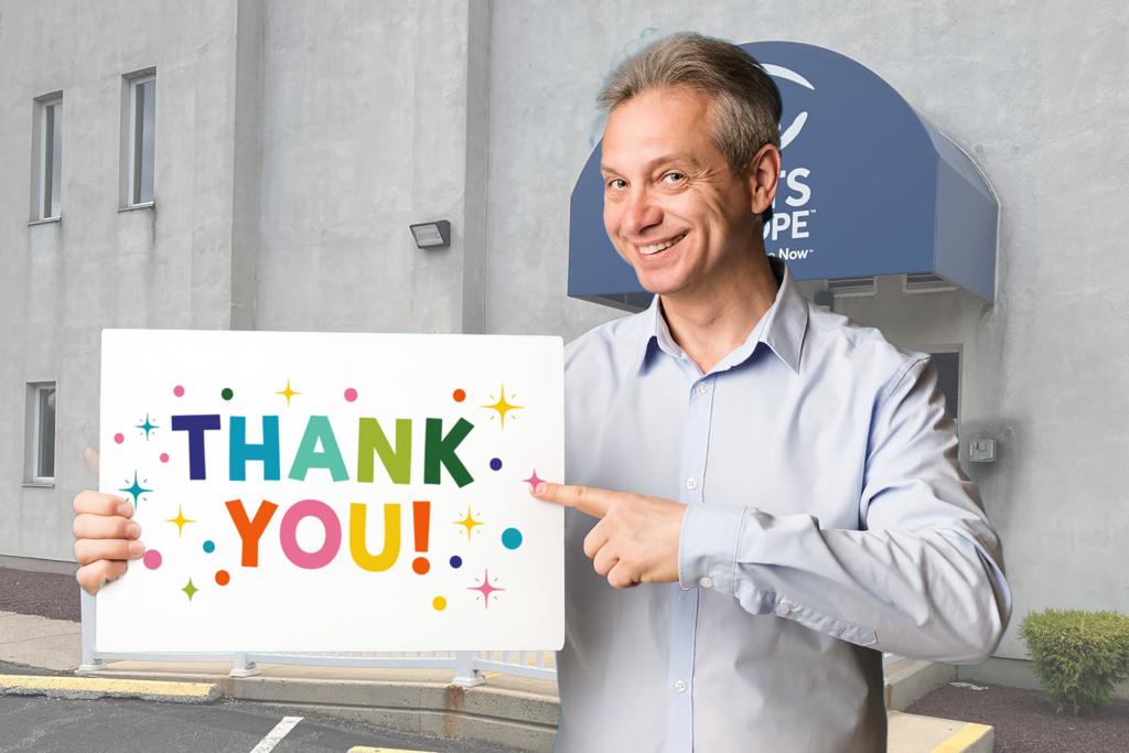 A smiling man in a light blue shirt holds a colorful "THANK YOU" sign with stars and dots while standing outside Sights for Hope’s Lehigh Valley Services Center.