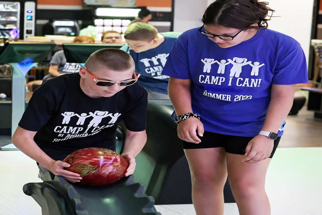 A boy wearing sunglasses and a "Camp I Can!" t-shirt places a bowling ball on a ramp, assisted by a smiling girl in a similar shirt, at a bowling alley
