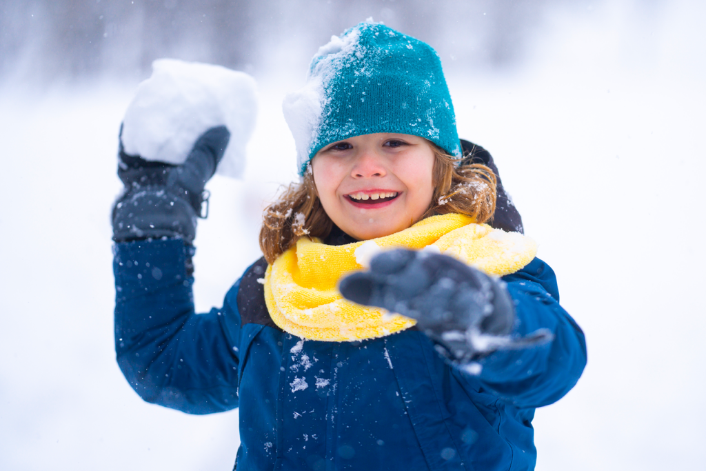 A child in a teal beanie and yellow scarf prepares to throw a snowball while smiling widely in a snowy landscape