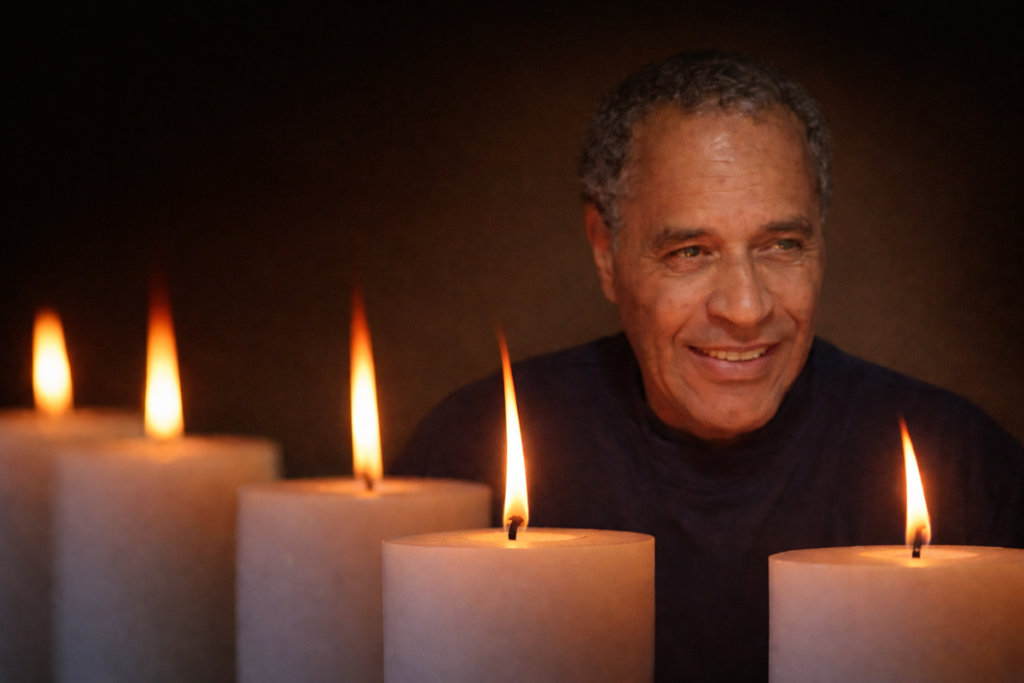 A man smiling warmly is surrounded by glowing candles in a dimly lit setting, creating a serene and contemplative ambiance.