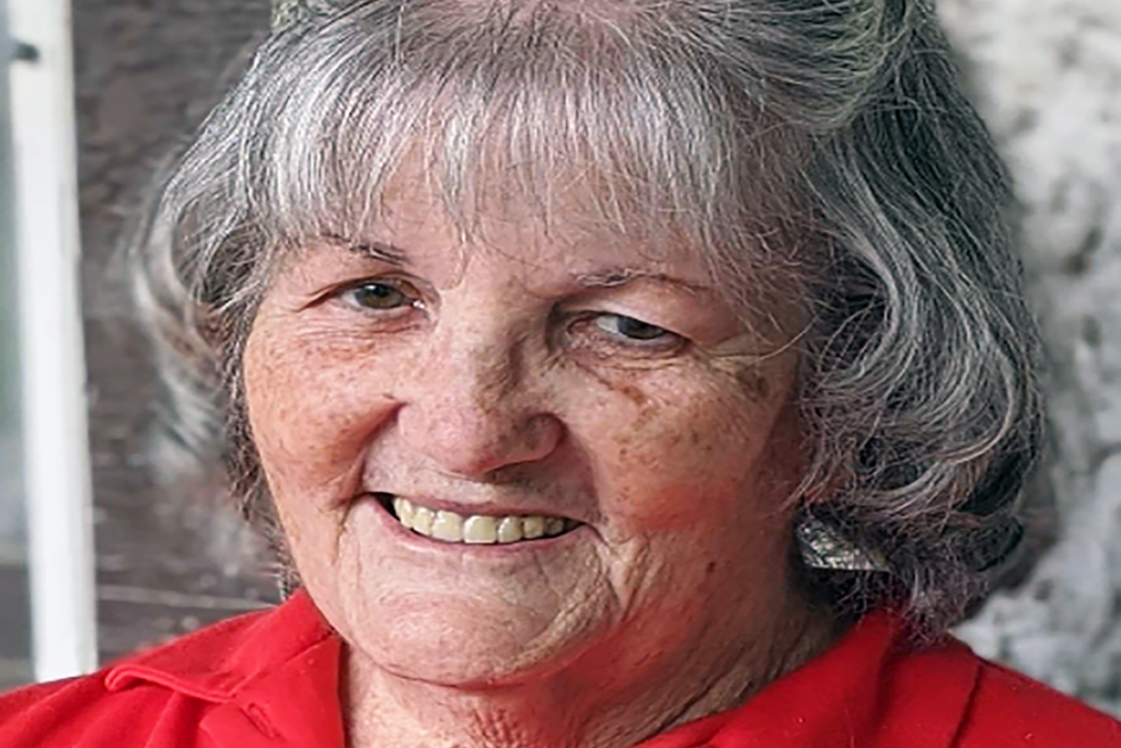 Older woman with gray hair and a warm smile, wearing a red shirt. She exudes a friendly demeanor. The background is softly blurred.