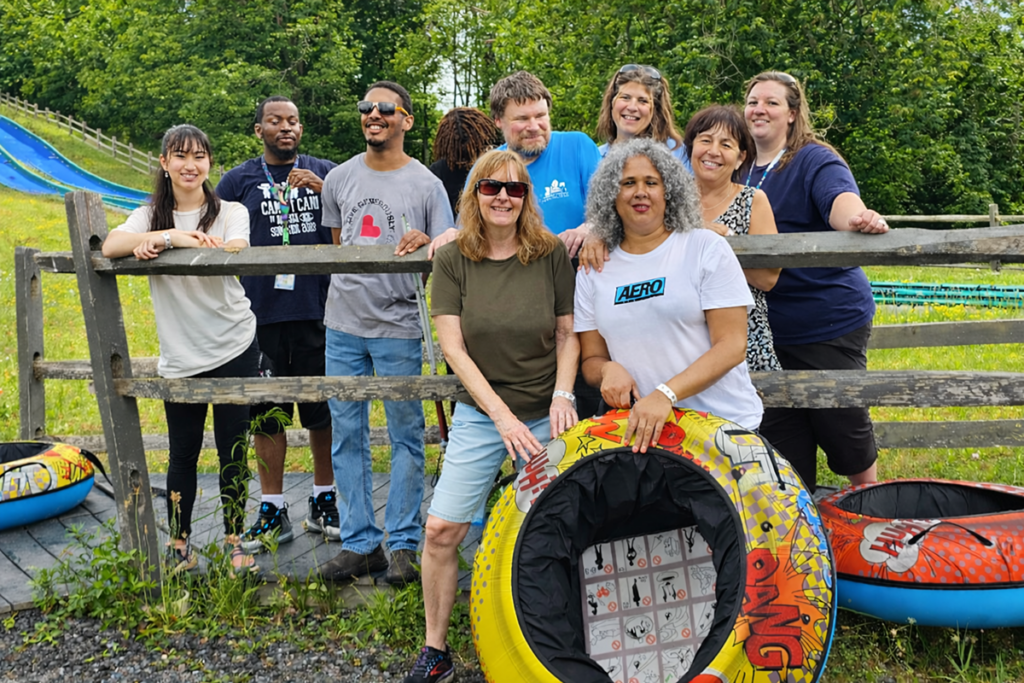 A group of nine adults poses happily outdoors near a wooden fence. Bright green grass and tubing equipment are visible, suggesting a fun, relaxed day of outdoor activities.