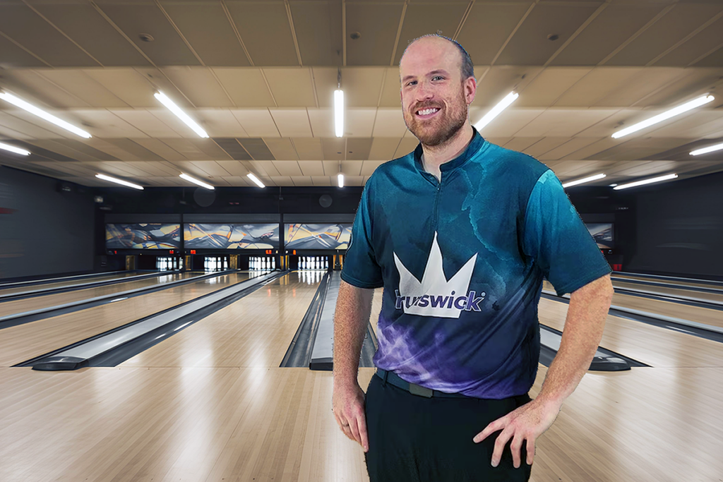 A smiling man in a bowling alley wears a blue and purple Brunswick shirt and stands confidently with with his hands on his hips