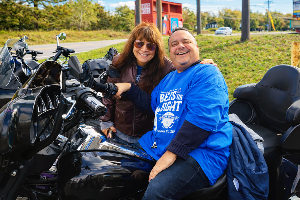 A woman in sunglasses stands beside and a man in a bright blue Sights for Hope shirt, who is sitting on a black motorcycle.