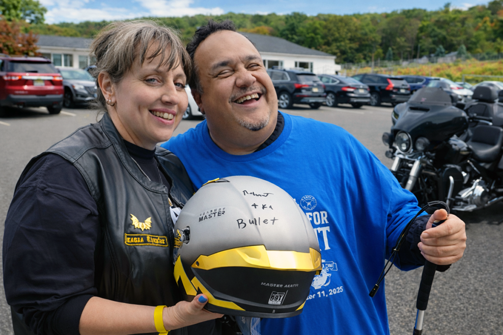 A joyful man and woman stand together, both smiling. The man wears a blue shirt, holding a white cane, and the woman wears a black leather vest, holding a helmet. Two motorcycles and several cars are parked in the background. The scene is outdoors with trees visible.